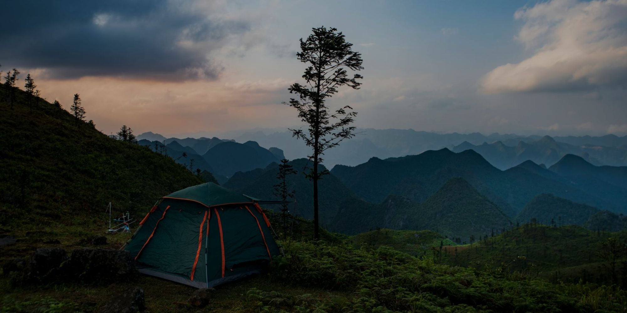 Camping tent on a hill with a tree and mountain range in the background during sunset.